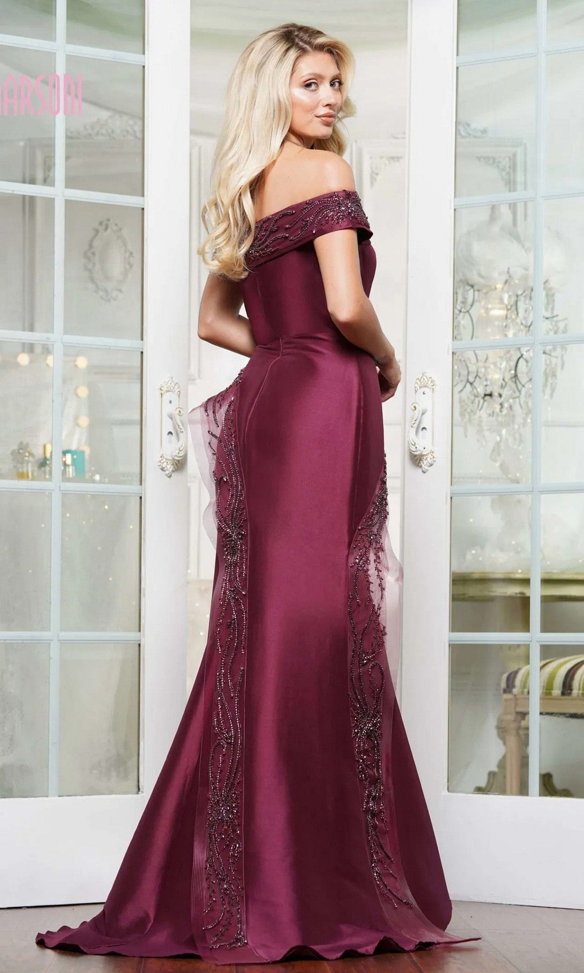 A woman stands indoors, facing away from the camera, wearing the Marsoni MV1332 long formal dress—an elegant burgundy off-shoulder mermaid gown with beaded embellishments in luxurious Mikado fabric.